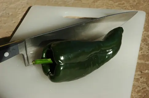 dark green poblano pepper with stem on a white cutting board beside a chef's knife, ready for preparation