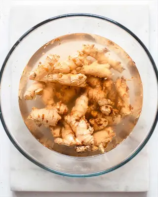 fresh ginger rhizomes soaking in water inside a clear glass bowl on a marble surface