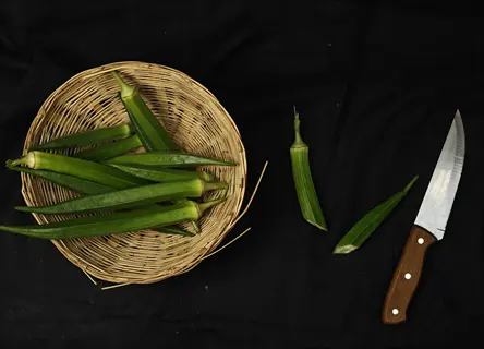 fresh okra pods in a woven basket with a knife and cut pieces on a black background, likely jambalaya okra