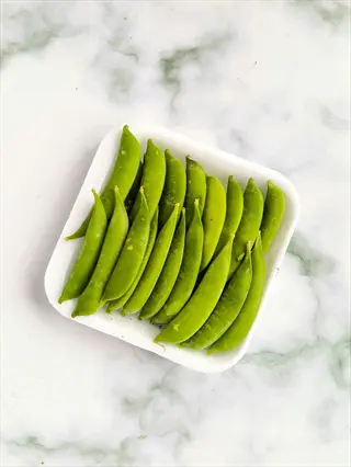 fresh shelling peas green pods (similar to wando variety) arranged in a white tray on marble surface