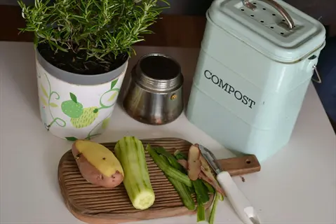 kitchen scraps including cucumber peels and potato on a cutting board next to a compost bin
