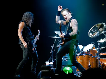 metallica band members performing live on stage with electric guitars and drum kit under concert lighting