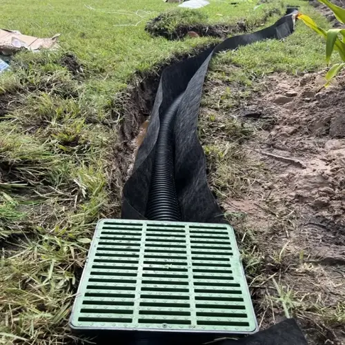 yard drainage french drain installation with green slatted cover, black perforated pipe in fabric-lined trench, surrounded by grass and dirt